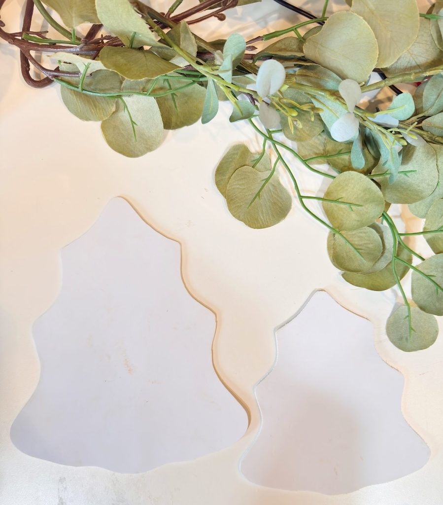 Two blank, white, tree-shaped objects are placed on a light surface with green, leafy branches arranged above them.