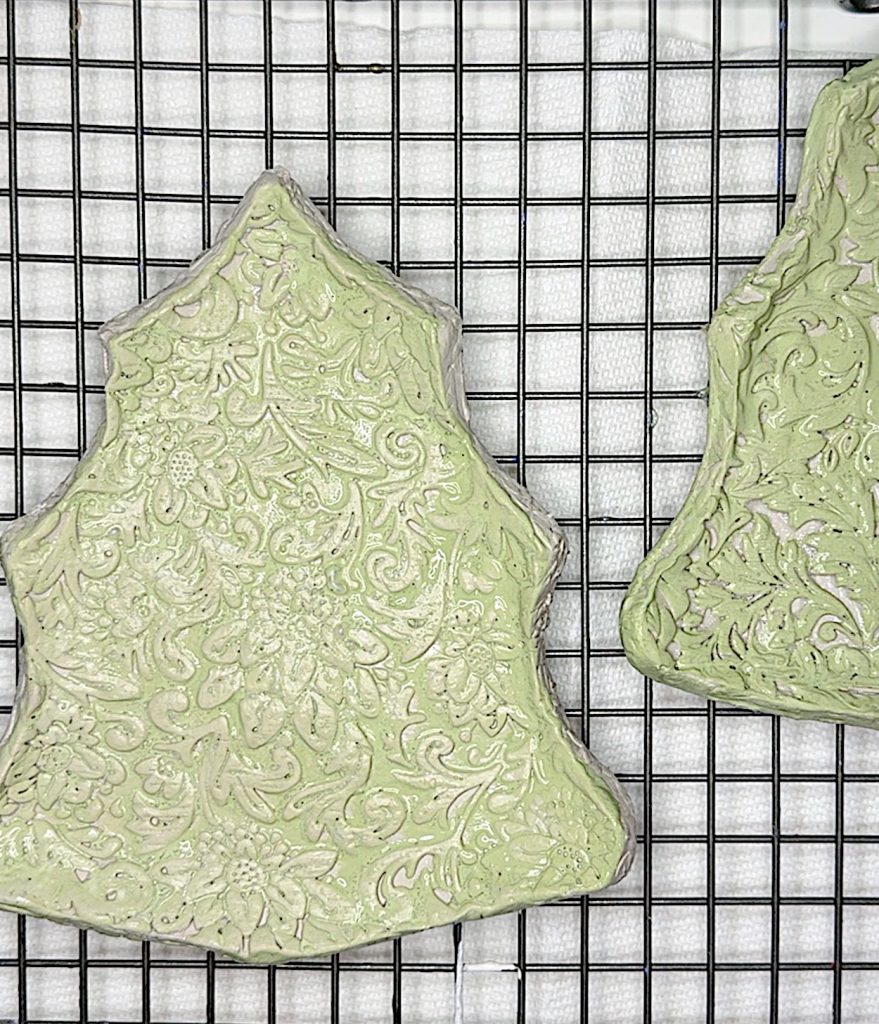 Two light green, tree-shaped cookies with intricate floral patterns are cooling on a black wire rack over a white textured surface.