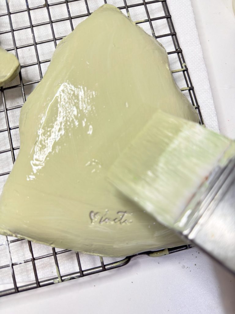 A pastry brush applies a light green glaze to a heart-shaped baked item on a wire rack, with the word "matcha" visible on the surface.