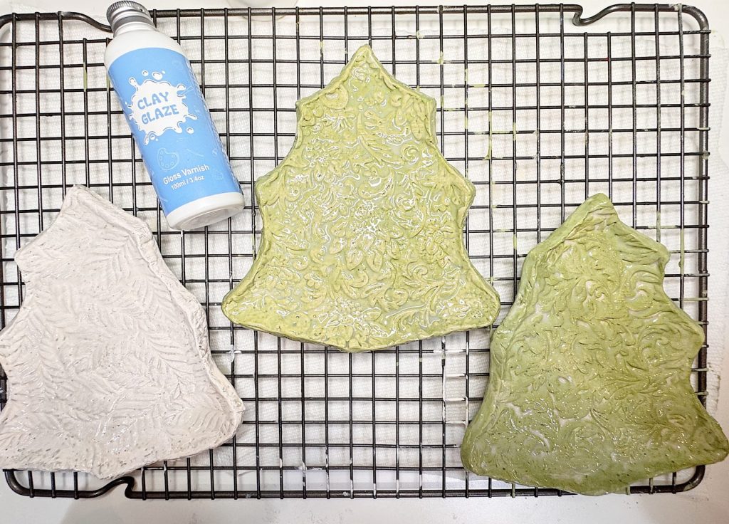 Three textured clay pieces shaped like Christmas trees are drying on a cooling rack next to a bottle labeled "Clay Glaze - Glossy Finish.