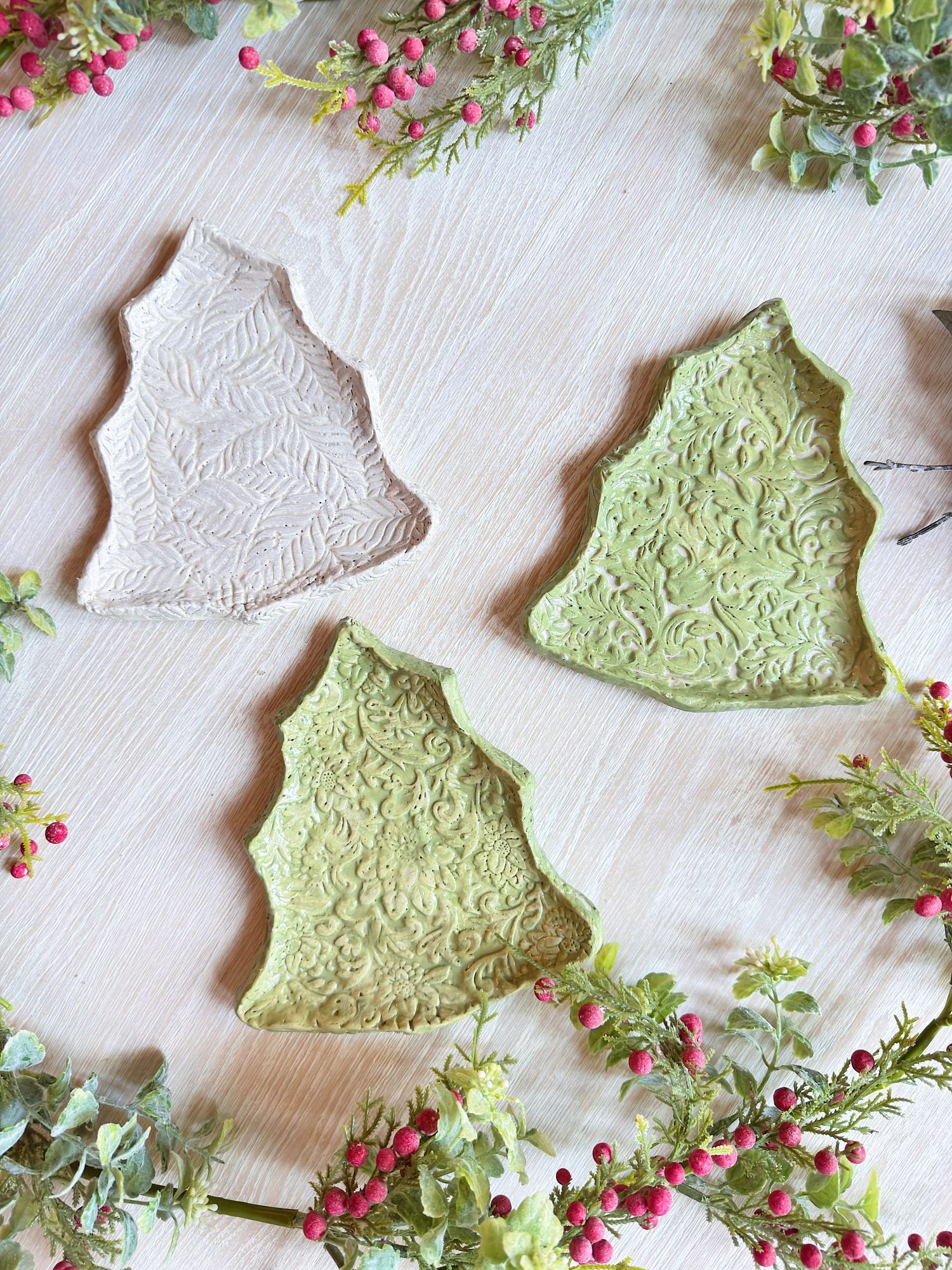 Three ceramic trays shaped like Christmas trees, two green and one beige, with textured patterns, are arranged on a light wooden surface surrounded by greenery and red berries.