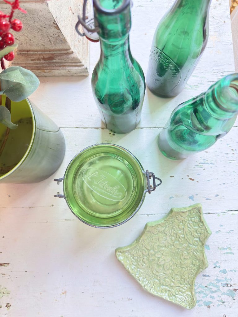 Three green glass bottles, a glass jar with green liquid, a green leafy plant in a pot, and a green ceramic dish shaped like a Christmas tree on a white wooden surface.