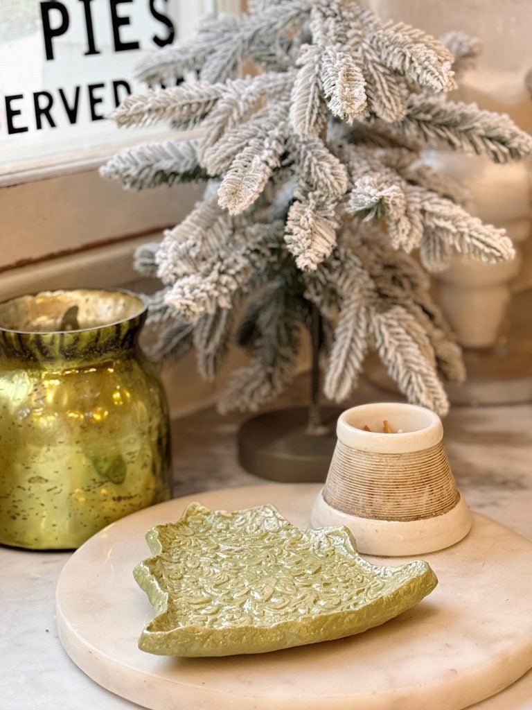 A small frosted artificial pine tree, a green decorative dish, a gold candle holder, and a beige candle are arranged on a marble surface.