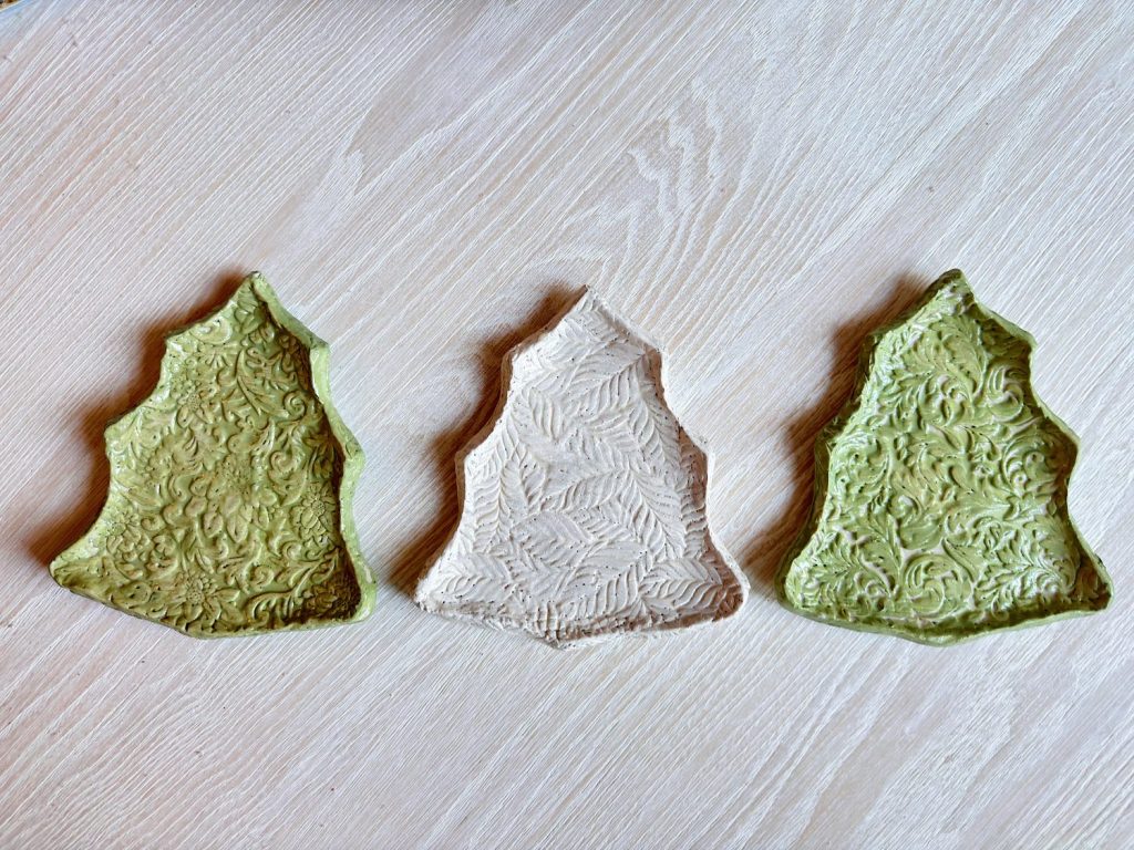 Three textured ceramic plates shaped like Christmas trees are arranged in a row on a light wooden surface; the two outer plates are green and the center plate is off-white.
