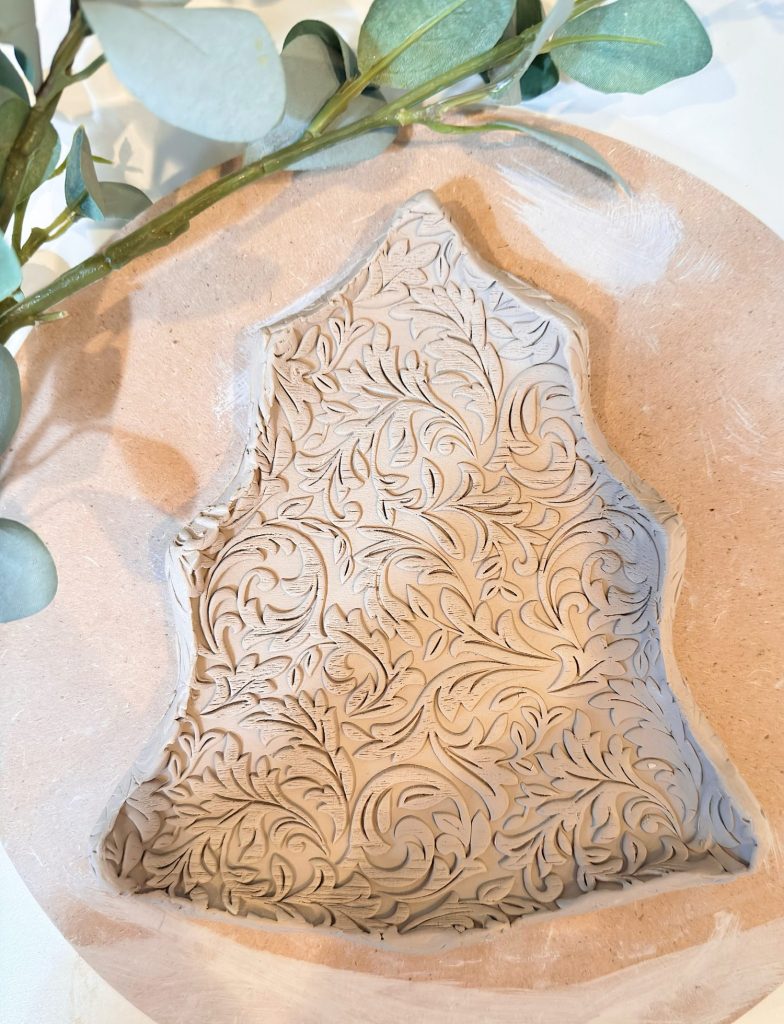 A clay piece shaped like a Christmas tree with intricate floral patterns is displayed on a round wooden board next to green leaves.
