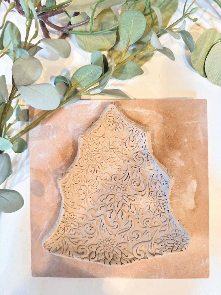 A clay piece shaped like a Christmas tree with intricate floral and leaf patterns sits on a square slab; eucalyptus branches are visible in the background.