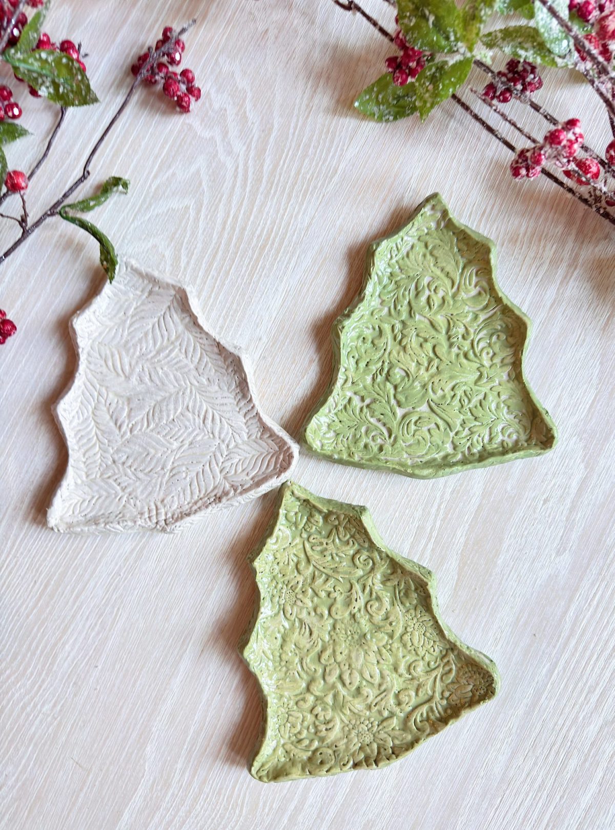 Three ceramic plates shaped like Christmas trees, two green and one white, with textured patterns, are arranged on a light wooden surface next to festive berry and leaf decorations.