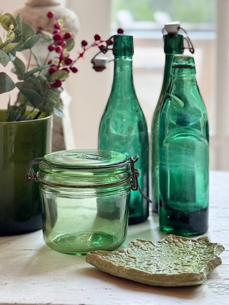 Green glass jar, two green glass bottles, a green vase with leaves and berries, and a textured green ceramic dish displayed on a light surface.