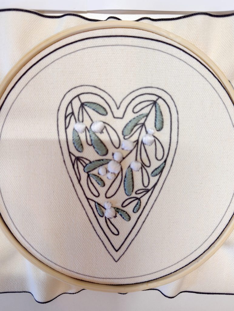 An embroidery hoop displays a heart-shaped design with green leaves and white flowers stitched on cream fabric.
