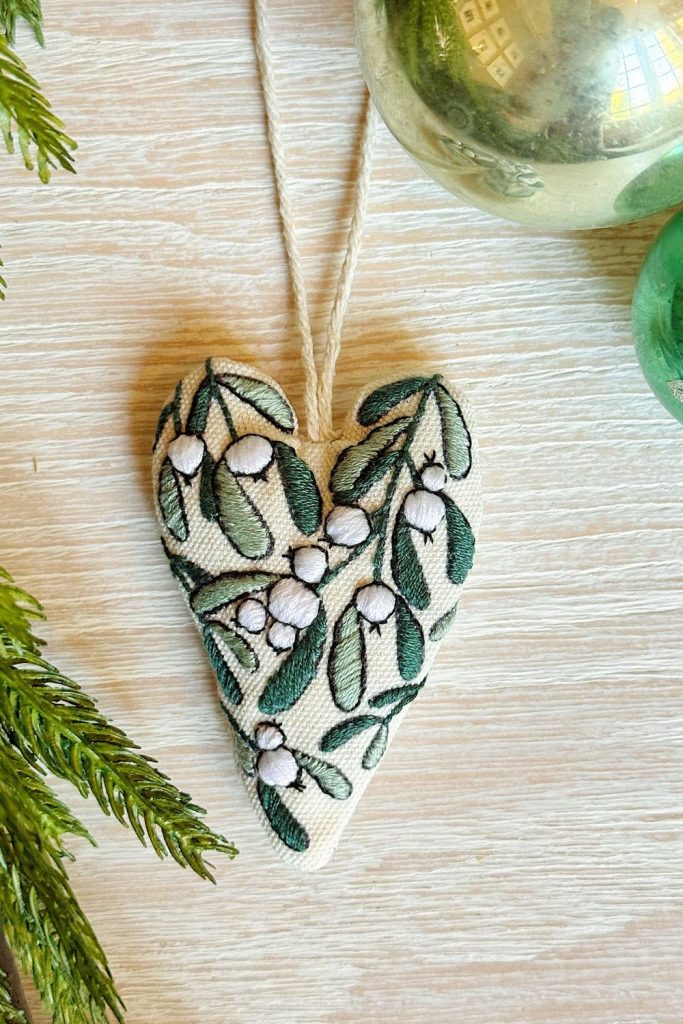 A heart-shaped fabric ornament with green leaves and white berries embroidery, lying on a light wood surface near green and gold holiday decorations.
