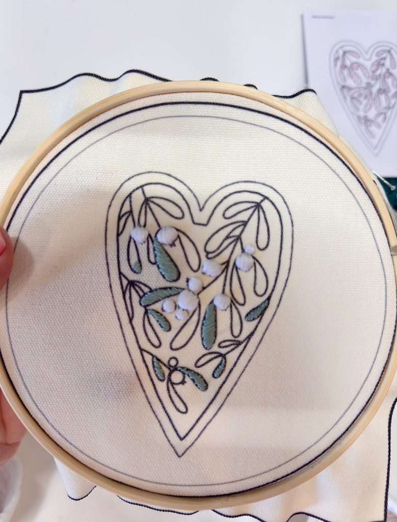A hand holds an embroidery hoop with fabric, displaying a heart-shaped outline and partially completed leaf and berry stitching inside the heart.