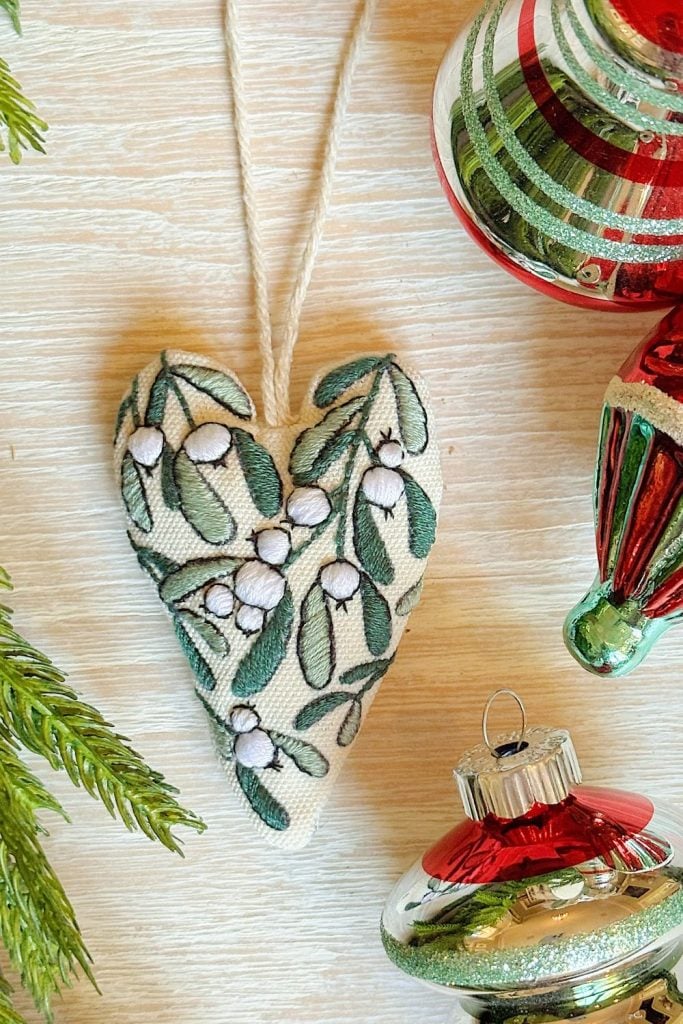 A fabric heart ornament with embroidered mistletoe is shown next to green pine needles and red and green striped Christmas baubles on a wooden surface.