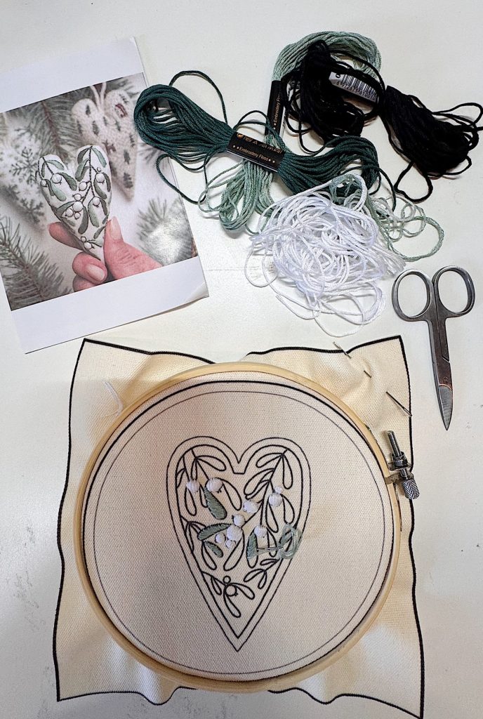 Embroidery kit with a heart-shaped pattern, embroidery floss, scissors, and a reference photo displayed on a table.