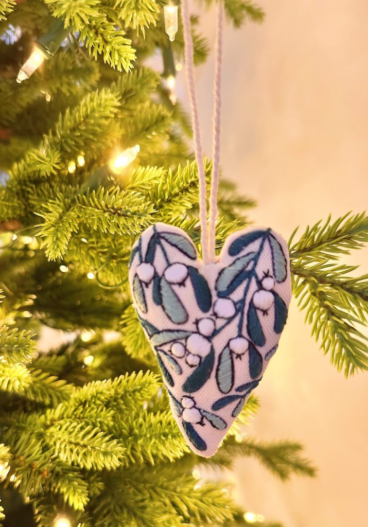 A heart-shaped ornament with a white and green leaf pattern hangs on a Christmas tree branch with warm white lights.