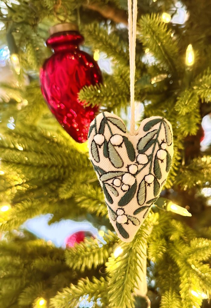 A fabric heart ornament with green leaves and white berries hangs on a Christmas tree next to a red glass ornament, surrounded by warm white lights.