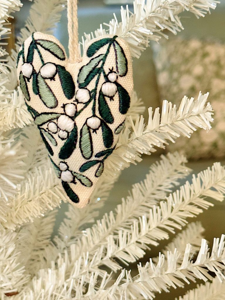 A white artificial Christmas tree decorated with a fabric heart ornament embroidered with green leaves and white berries.