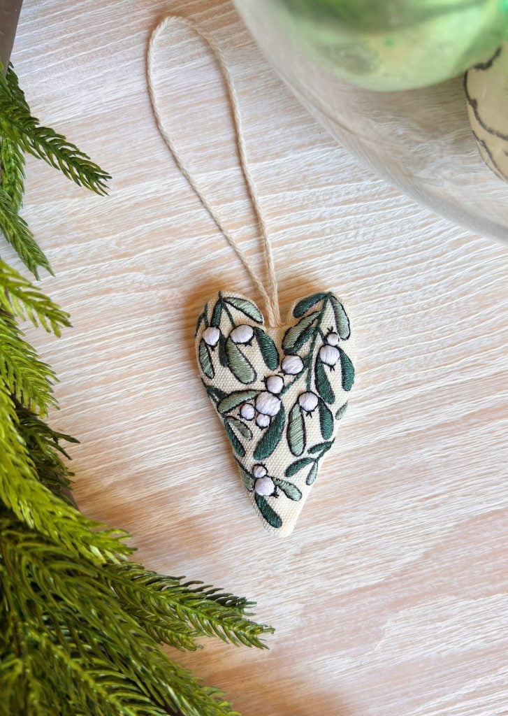 A heart-shaped fabric ornament with embroidered green leaves and white flowers lies on a light wooden surface next to a sprig of greenery.
