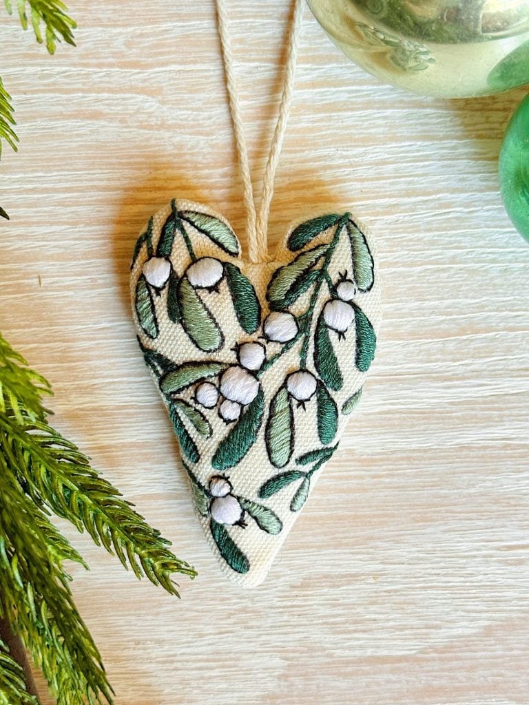 A heart-shaped fabric ornament with embroidered green leaves and white berries, hanging from a string on a light wooden surface. Greenery is partially visible at the left and top right corners.