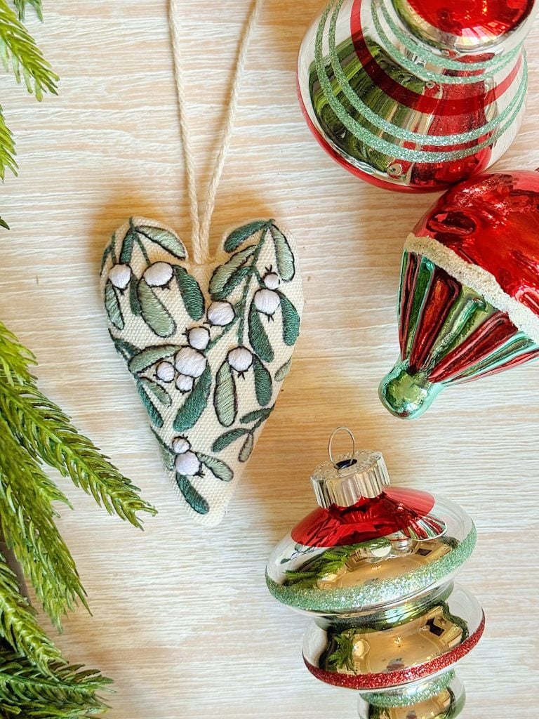 A fabric heart ornament with a botanical design lies next to three red and green glass Christmas ornaments and some evergreen branches on a light wooden surface.