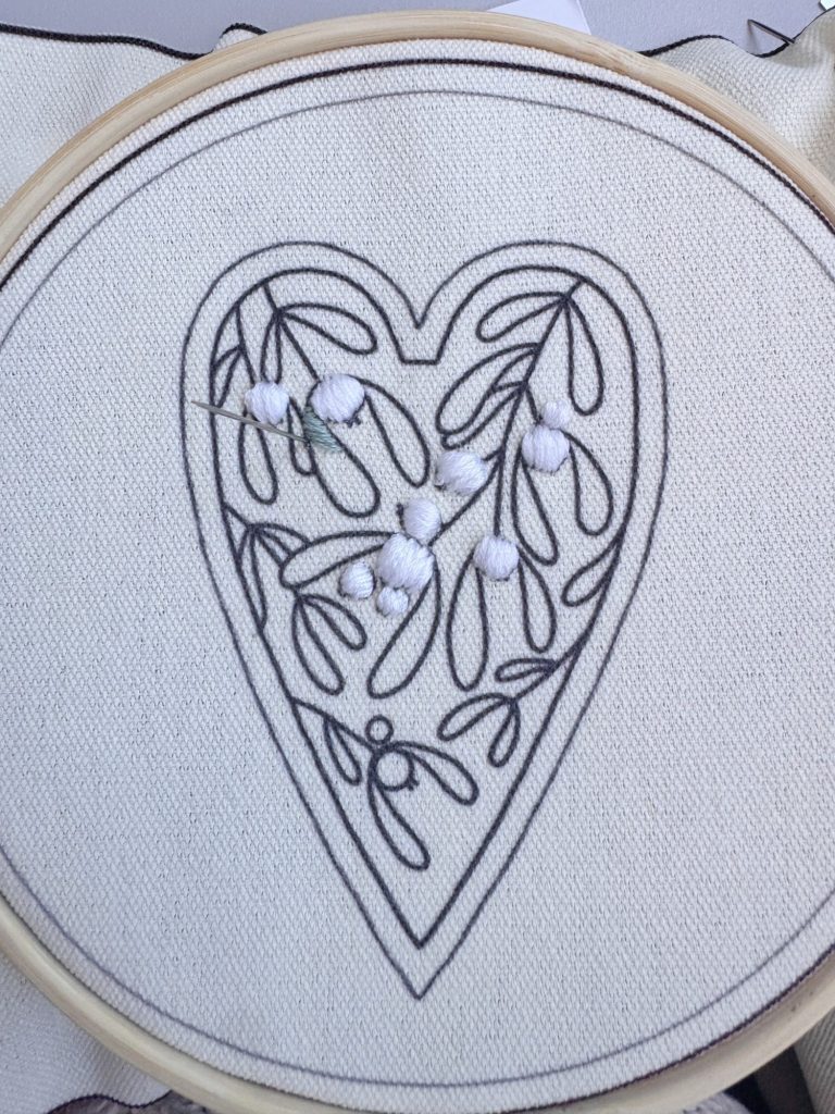 Black outline embroidery of a heart with leaf and berry design on white fabric, some berries partially filled with white thread, held in an embroidery hoop.