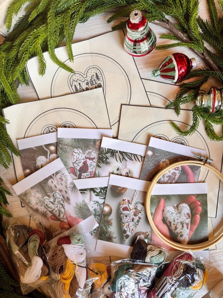 Embroidery supplies, hoop, thread, and printed heart designs are arranged on a table with holiday ornaments and greenery in the background.