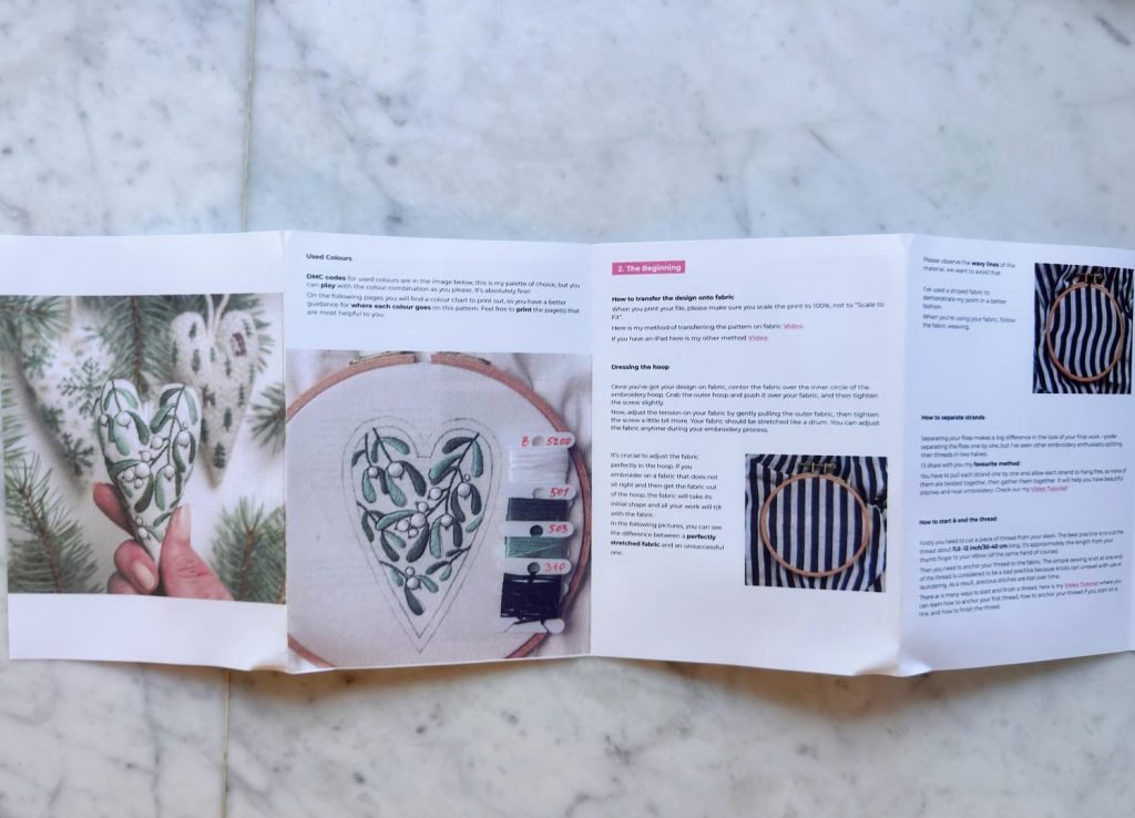 A folded instruction booklet on a marble surface shows photos and text about an embroidery project featuring a heart-shaped leaf design.
