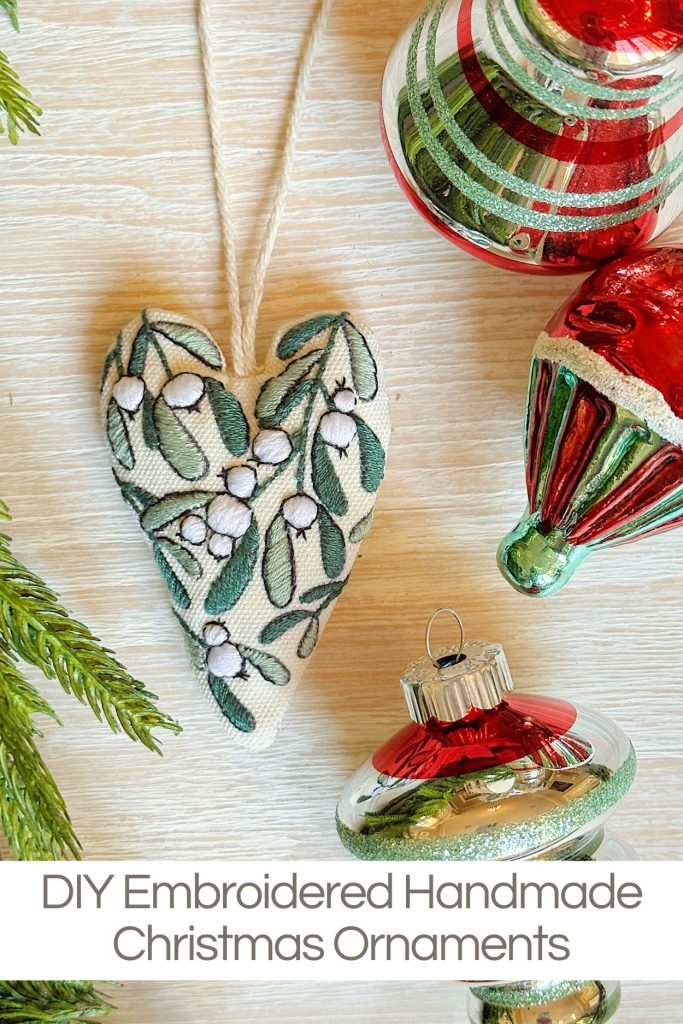 Three Christmas ornaments, including a DIY embroidered heart with green leaves and red and green glass baubles, are displayed on a light surface with pine branches nearby.
