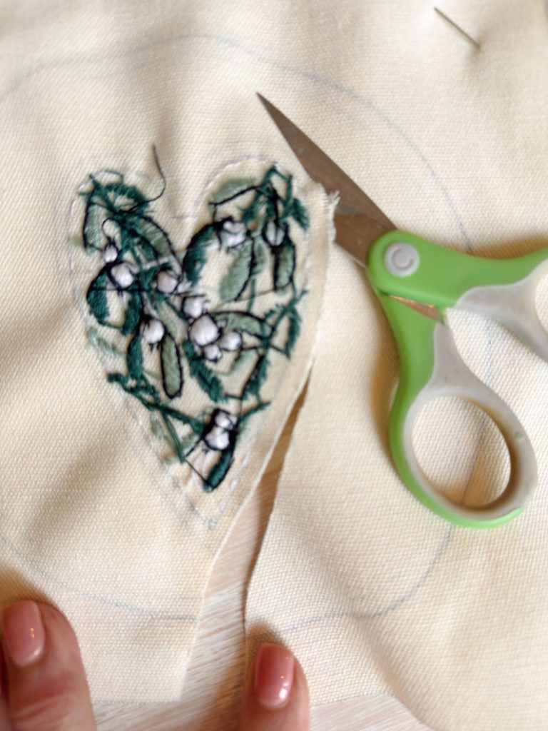 A piece of fabric with a heart-shaped floral embroidery design is being cut out with green-handled scissors; a hand is visible in the corner.