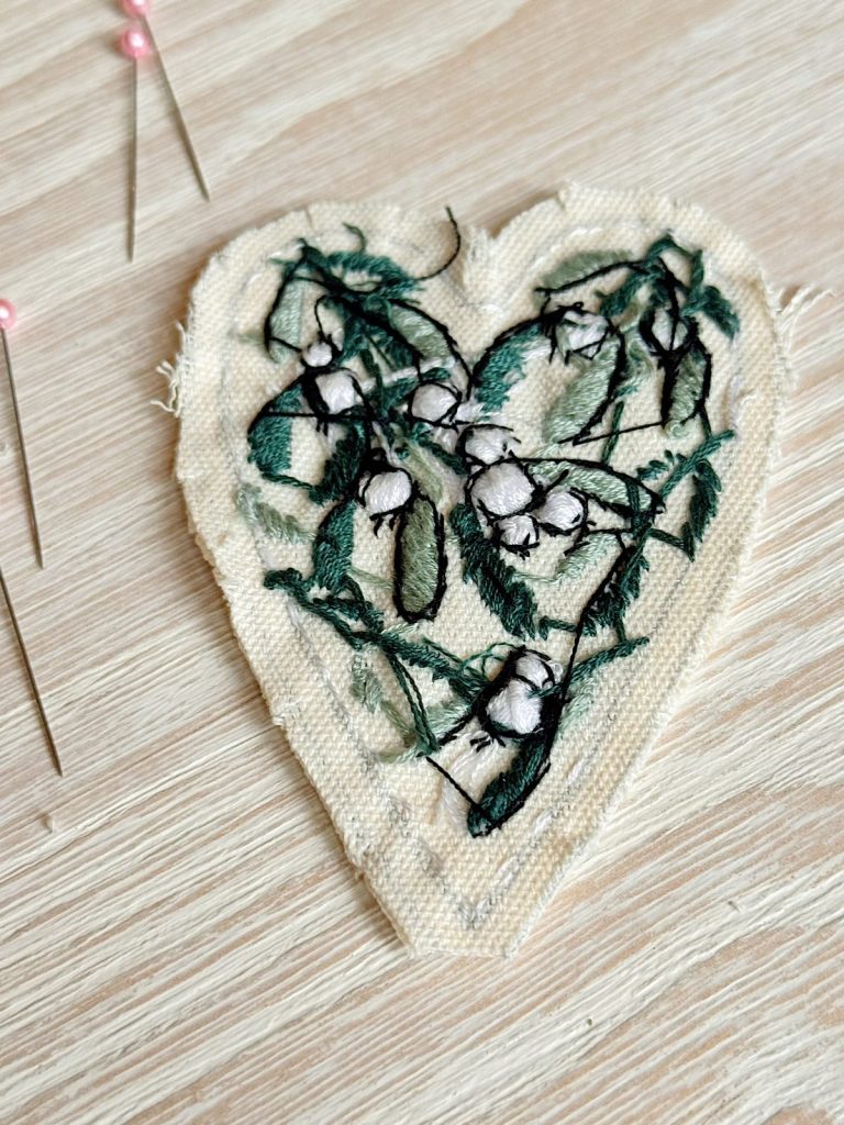 A heart-shaped piece of fabric with green and white floral embroidery, placed on a wooden surface next to sewing pins.