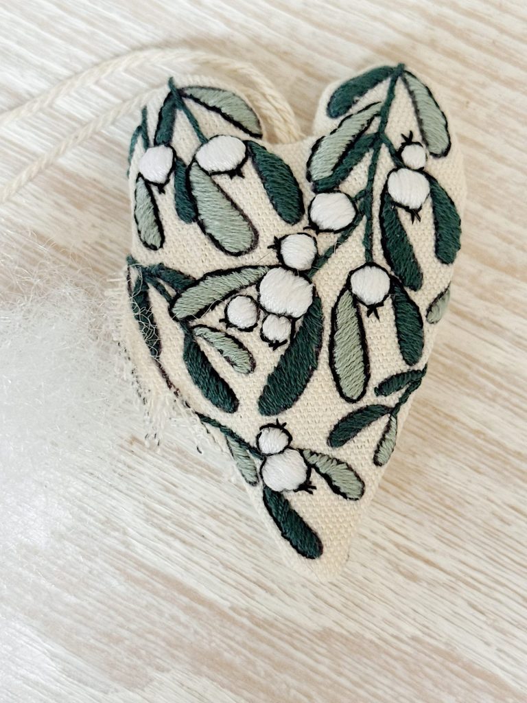 A fabric heart ornament embroidered with green leaves and white berries lies on a light wood surface, with some stuffing visible on the side.