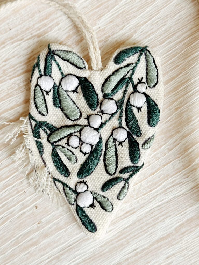 A fabric heart-shaped ornament with green and white embroidered leaves and berries, lying on a light wood surface.