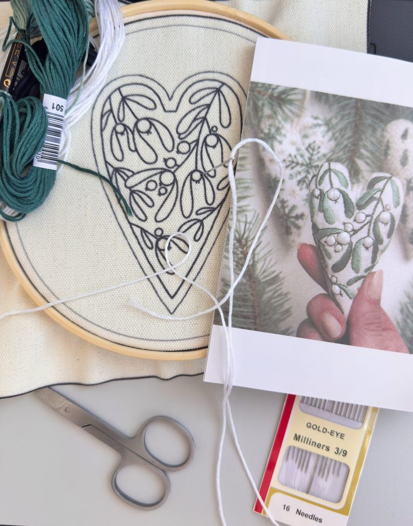 A hoop with fabric displays a heart-shaped embroidery outline beside thread, scissors, needles, and a reference photo of a finished embroidered heart.