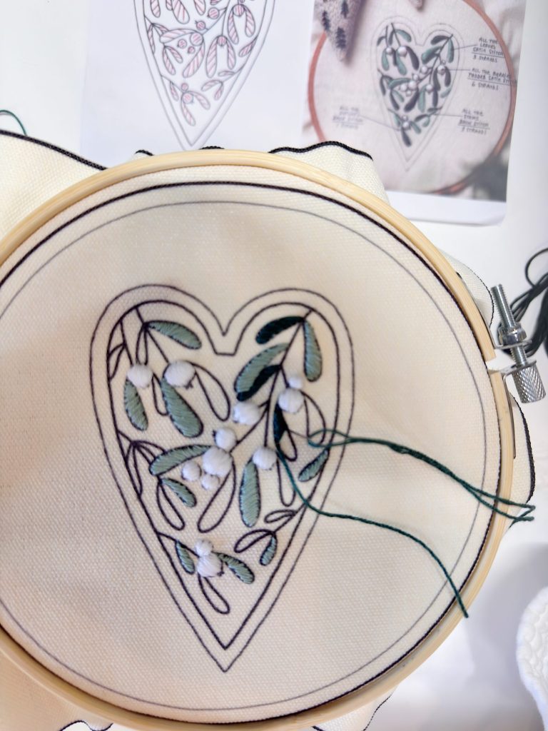 An embroidery hoop holds fabric with a heart-shaped, leafy design being stitched in green and white. A printed pattern reference is visible in the background.