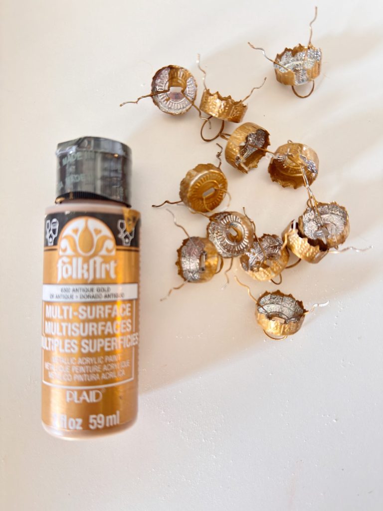 A bottle of gold multi-surface acrylic paint lies next to several metallic gold-painted bottle caps on a white surface.