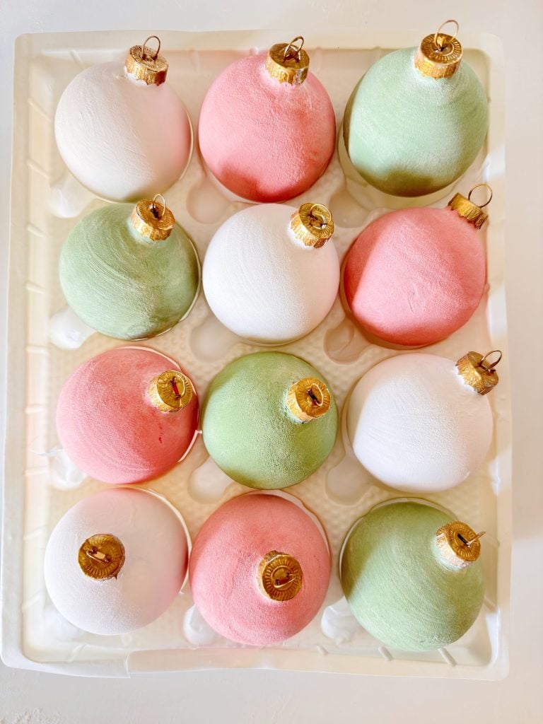 A tray holds twelve matte Christmas ornaments in pastel pink, green, and white, arranged in a neat grid.