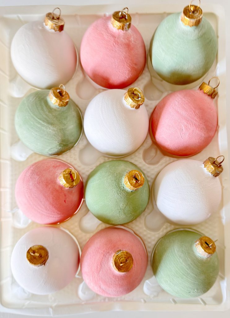 A tray containing twelve Christmas ornaments in pastel shades of pink, green, and white, arranged in a grid pattern.