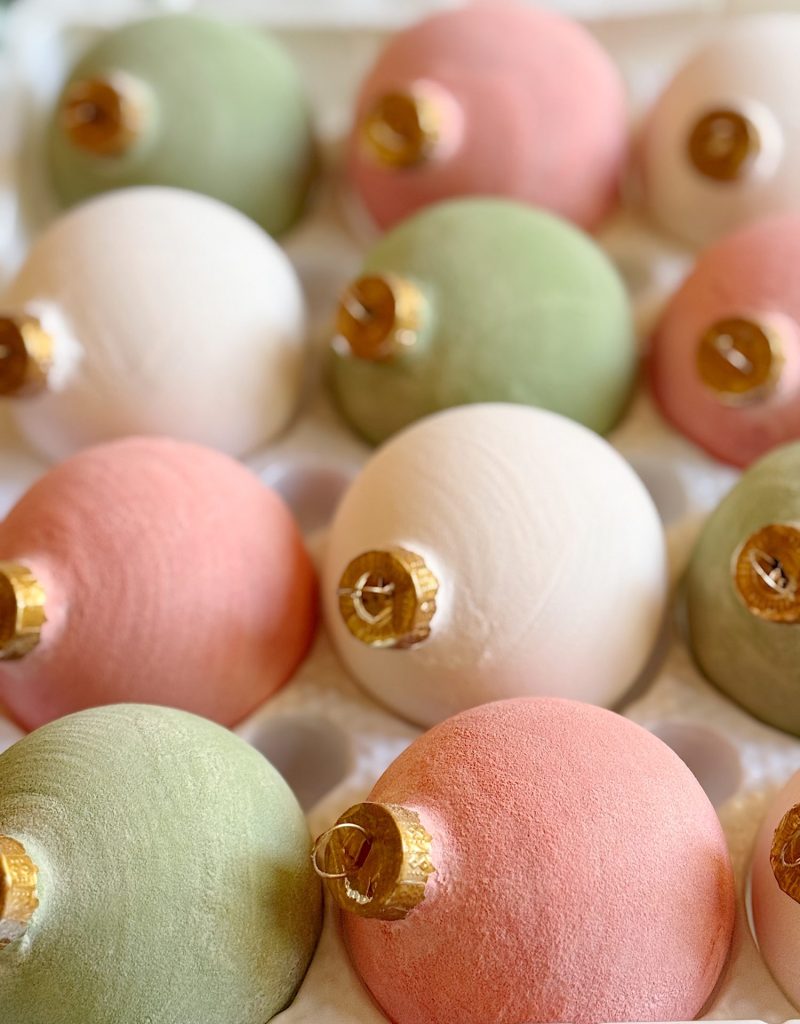 A close-up of pastel-colored Christmas ornaments in pink, green, and white, arranged closely together in a protective tray.