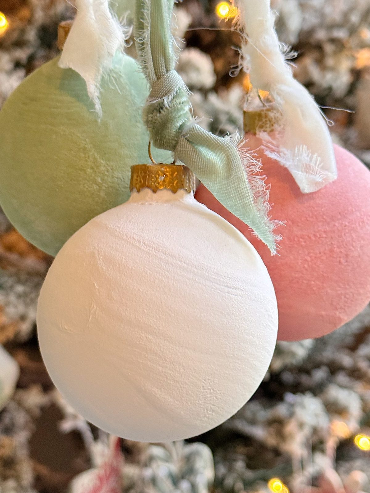 Three matte Christmas ornaments—white, light green, and pink—hang from frayed fabric ribbons in front of a blurred, decorated tree.