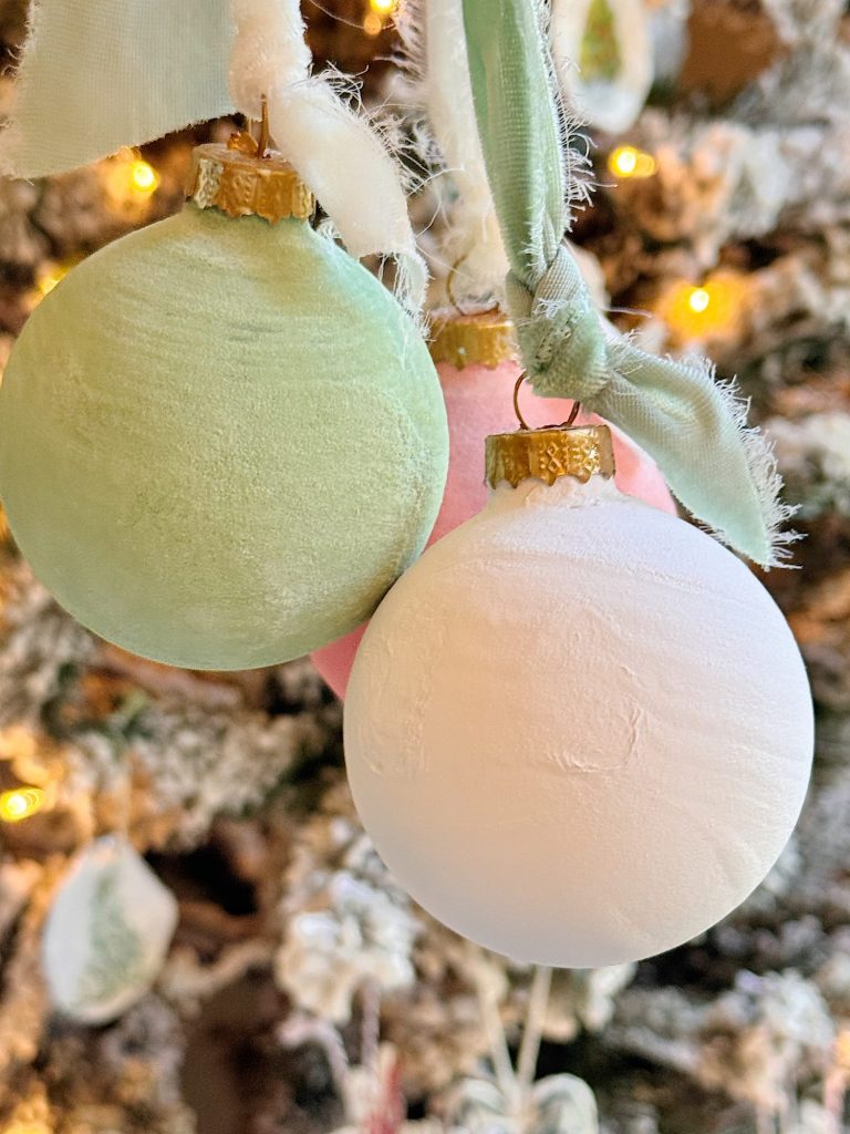 Three pastel-colored Christmas ornaments, including green, pink, and white, hang from ribbons in front of a decorated, lit Christmas tree.