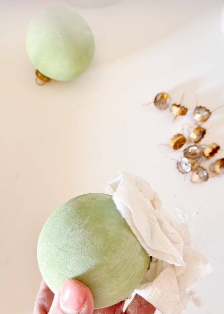 A hand wipes a green ornament with a tissue, with more green ornaments and gold ornament tops scattered on a white surface.