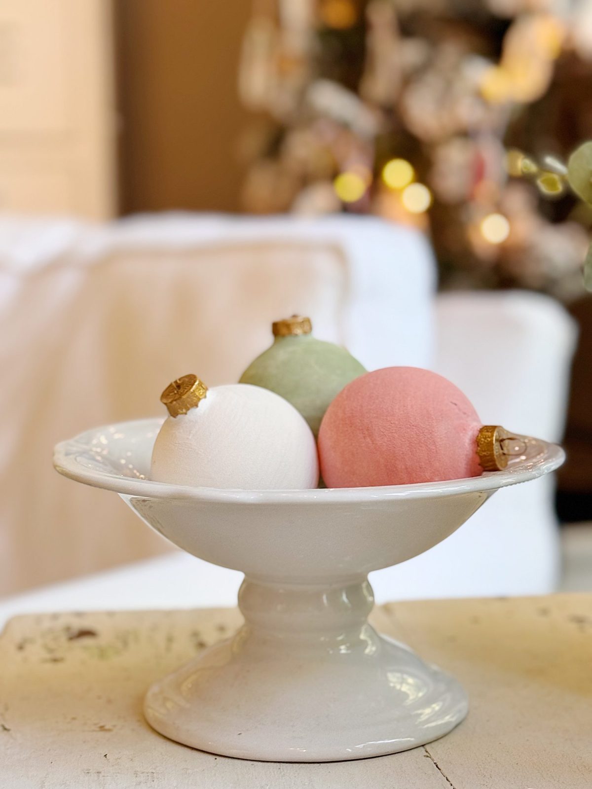 A white pedestal dish holds three pastel-colored ornaments—white, green, and pink—each with a gold cap, with a blurred background.