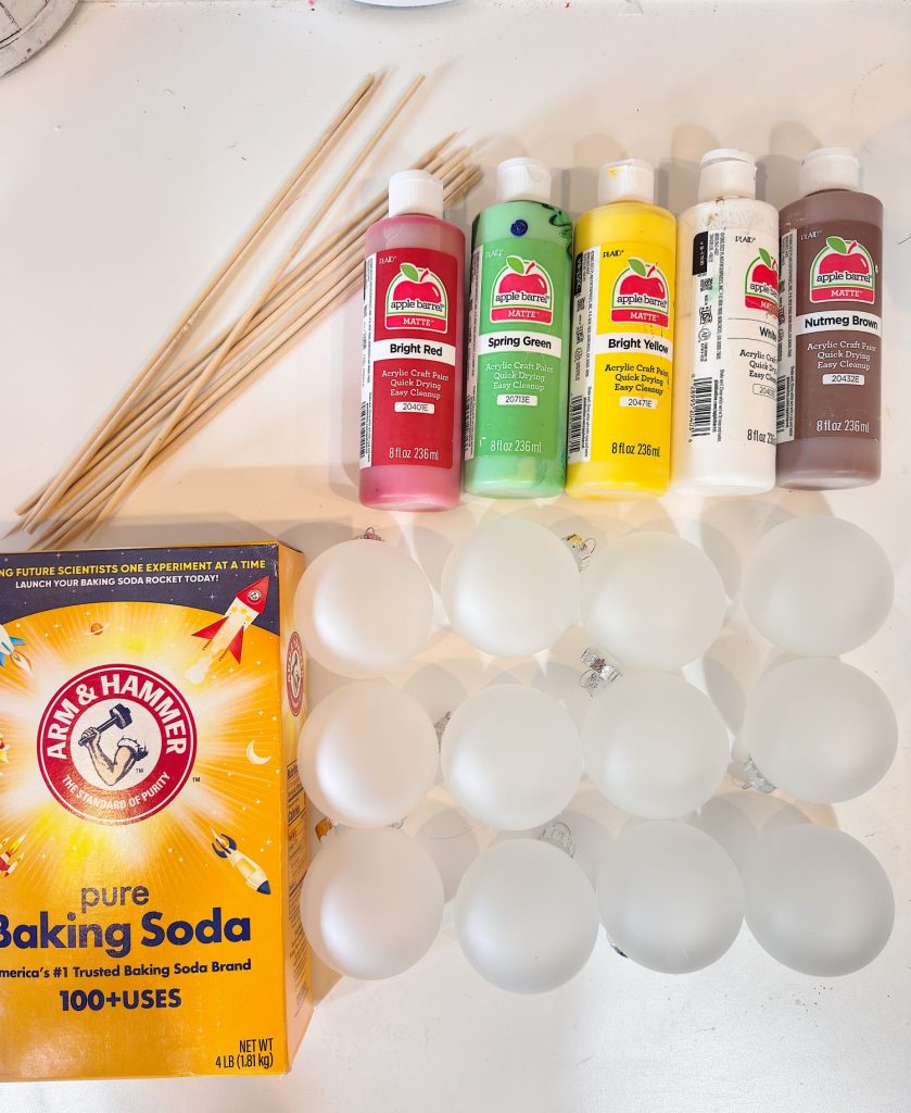 Art supplies on a white surface, including acrylic paint bottles, a box of baking soda, wooden skewers, and several clear plastic ornaments.