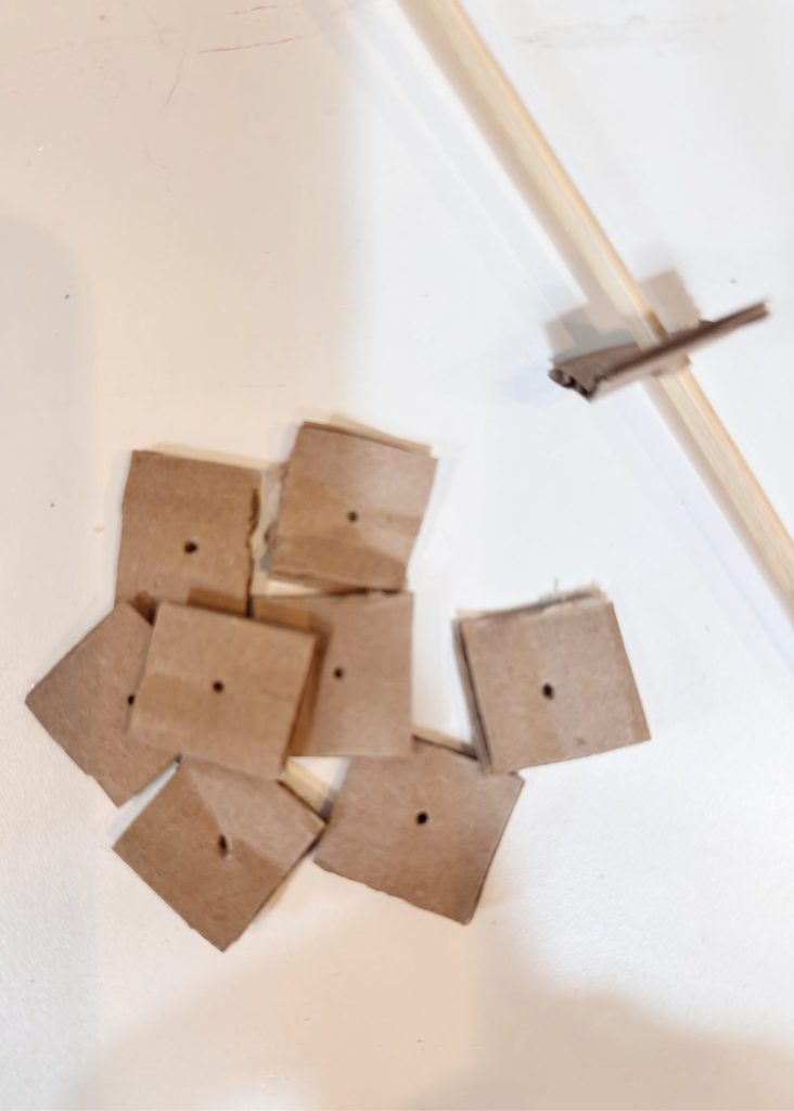 Eight small square cardboard pieces with center holes are scattered on a white surface next to a wooden stick with a piece of cardboard attached.
