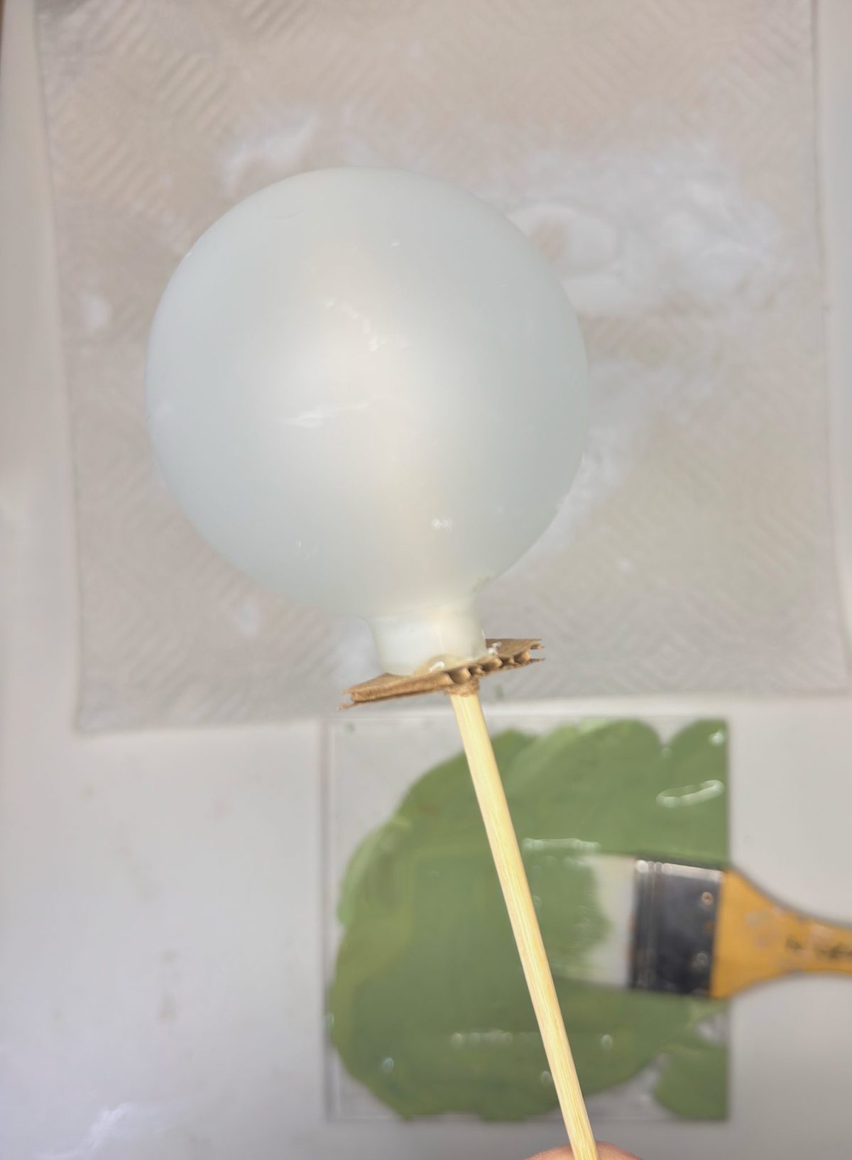 A white balloon is attached to a wooden skewer, held above a green painted surface with a paintbrush, and a paper towel is in the background.