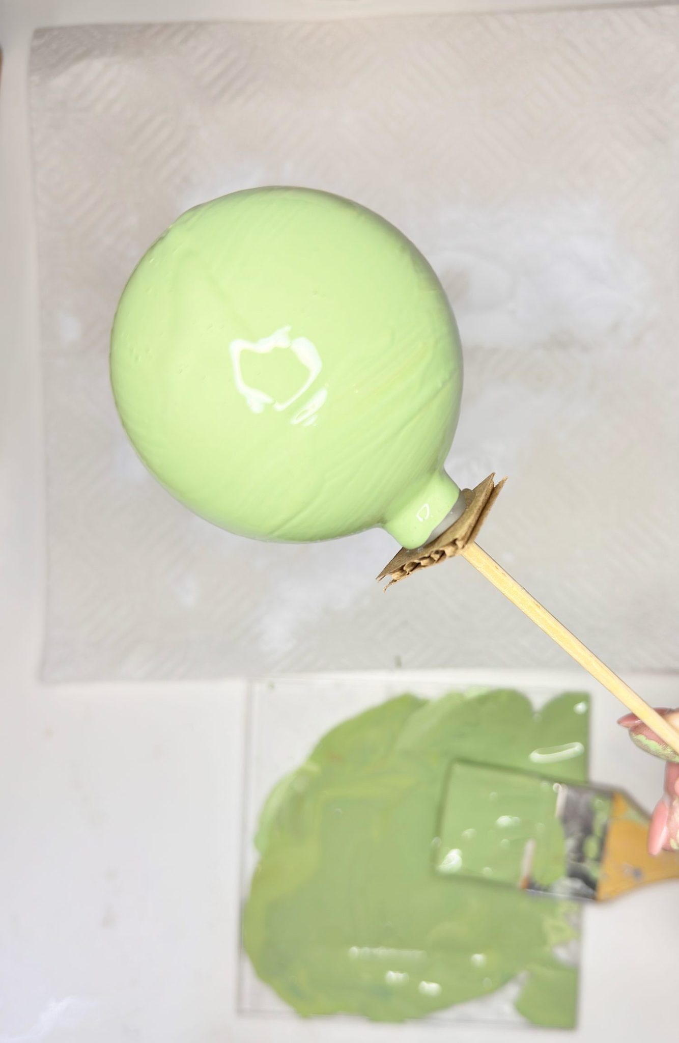 A hand holds a stick with a round balloon coated in light green paint above a tray containing more green paint.