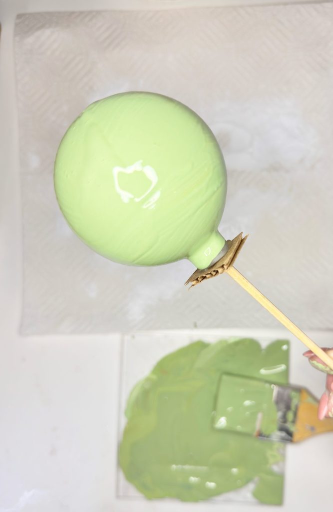 A hand holds a stick with a round balloon coated in light green paint above a tray containing more green paint.