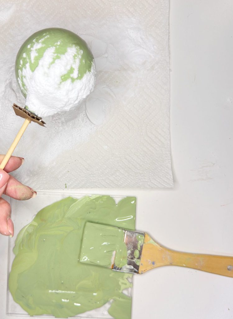 A hand holds a stick with a partially painted green sphere coated in white powder above a paper towel. Below are a paintbrush and a small tray with green paint.
