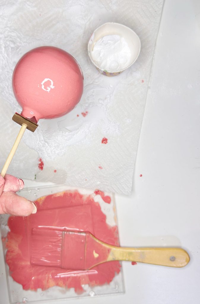 A hand holds a pink-painted round object on a stick above a napkin, with a paintbrush and pink paint on a plastic tray and a cup of white paint nearby.