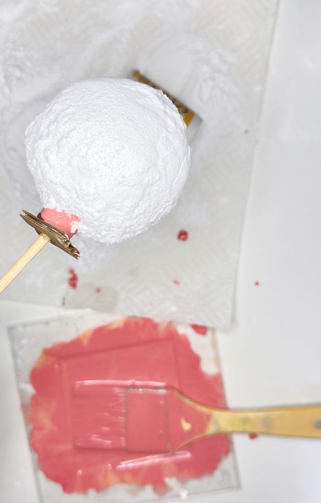 A white spherical object on a stick is held above a paper towel, with a brush and a tray of reddish-pink paint below.