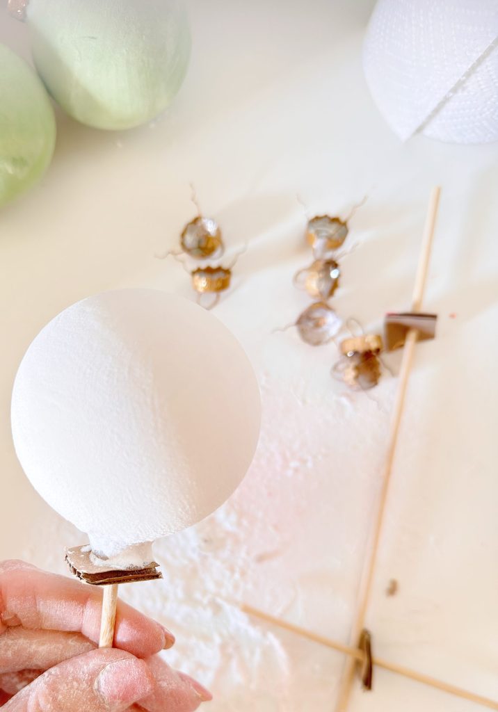 A hand holds a round white ornament on a stick, with paint powder on fingers. In the background are green ornaments and small decorative items on a white surface.